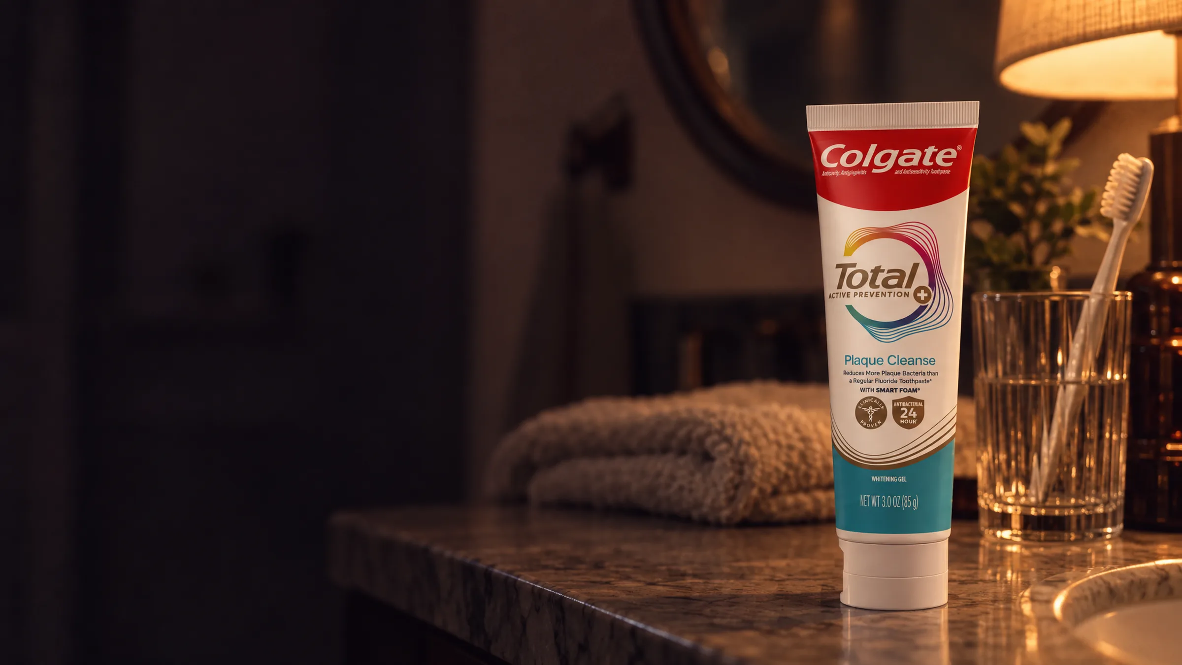 A tube of Colgate Total toothpaste on a bathroom counter at night, beside a glass with a toothbrush