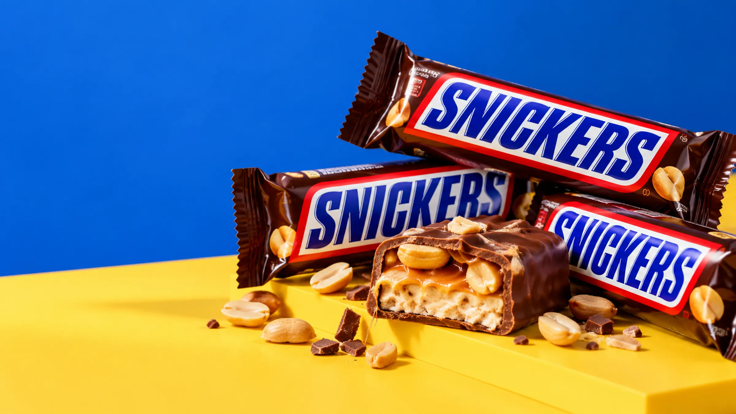 Several Snickers chocolate bars on a yellow surface with one bar open to show peanuts inside
